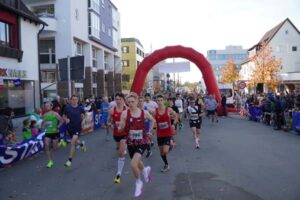Läuferinnen und Läufer des Hauptlaufs starten unter einem roten Bogen, Zuschauer säumen die Straße.