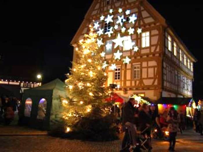 Beleuchteter Weihnachtsbaum und Marktstände vor einem Fachwerkhaus bei Nacht