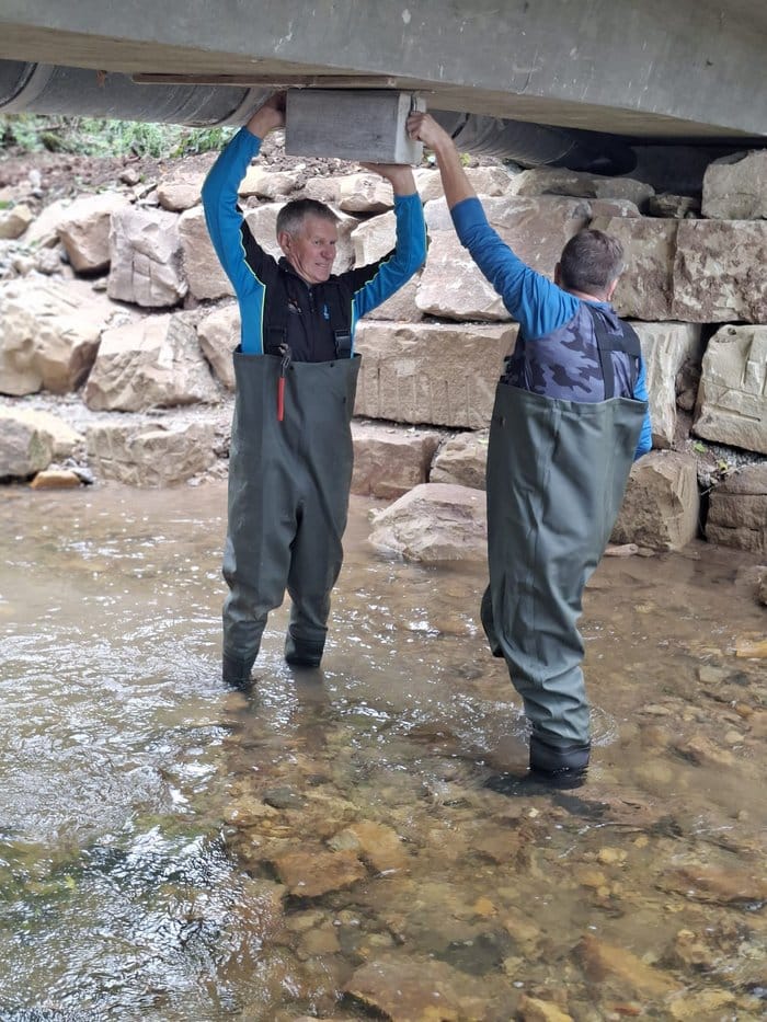 Zwei Helfer in Wathosen bringen unter einer Brücke einen Nistkasten an